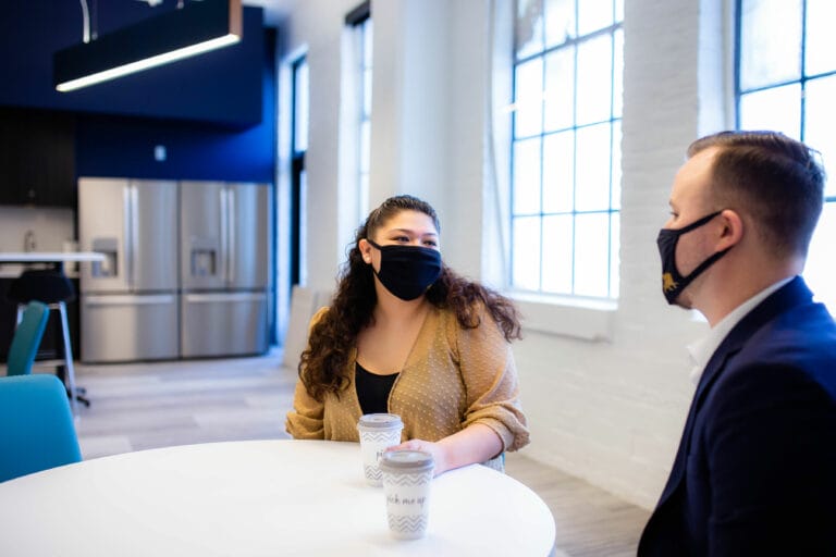 Young woman wearing a mask conversing in a breakroom with a man also wearing a black mask.