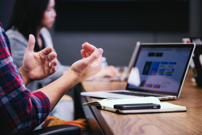 Image of a laptop on desk with a person working behind it. You can only see the person's hands and a blurred figure of somebody in the background.