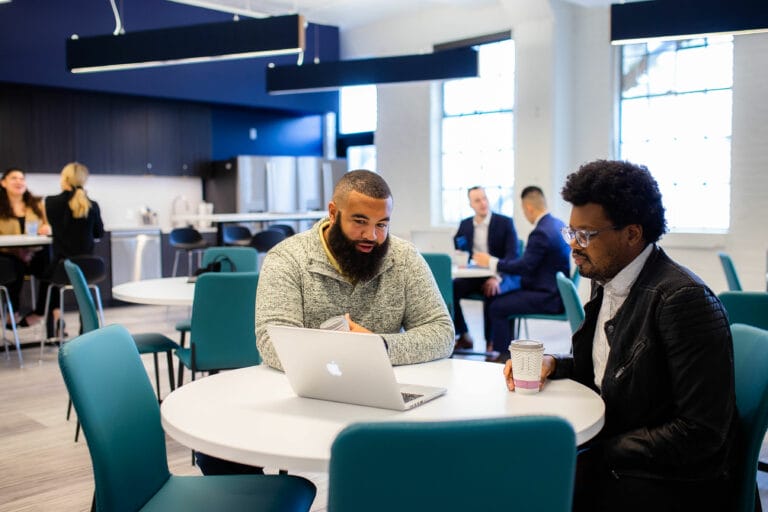 Image of two men sitting at a roudn table discussing something on the laptop in front of them.