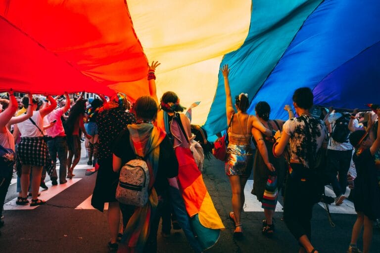 Group of parade participants marching under the gay pride flag. Photo provided by Unsplash.